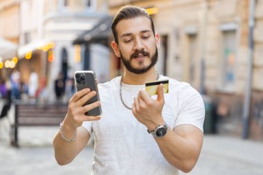 Genç adam para transfer ederken kredi kartı akıllı telefonu kullanıyor, online alışveriş yapıyor, yemek siparişi veriyor, otel odası ayırtıyor. Şehir güneşli caddelerde yürüyen bir turist.