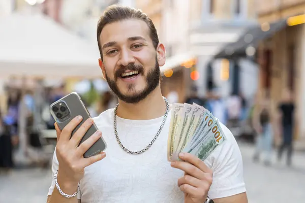 幸せな豊かな若い男がお金を数えるドル現金 スマートフォンの電卓アプリを使用して 贈り物や食品の配達をオンラインで注文する計画 ホテルの部屋を予約します 街の通りで髭男の観光客 屋外の町 — ストック写真
