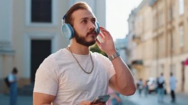 Neşeli, gevşemiş, sakallı, kablosuz kulaklıklı genç adam dışarıda dans eden enerjik disko rock 'n roll müziğini dinliyor. Şehir Güneşi Şehri arka planında yürüyen adam.