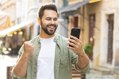 Kafkasyalı genç adam cep telefonuyla kutlama yapıyor. İyi haber kazan, piyango zaferi kazan, açık havada internetten bağış yap. Şehir caddesinde yürüyen mutlu yetişkin erkek turist. Kasaba yaşam tarzları