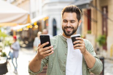 Mutlu gülümseyen adam akıllı telefon kullanıyor mesajlar yazıyor internet sosyal medya uygulamalarına göz atıyor internette sohbet ediyor sabahın tadını çıkarıyor sıcak kahve içmek için dışarı çıkıyor. Şehir caddesinde yürüyen bir adam.