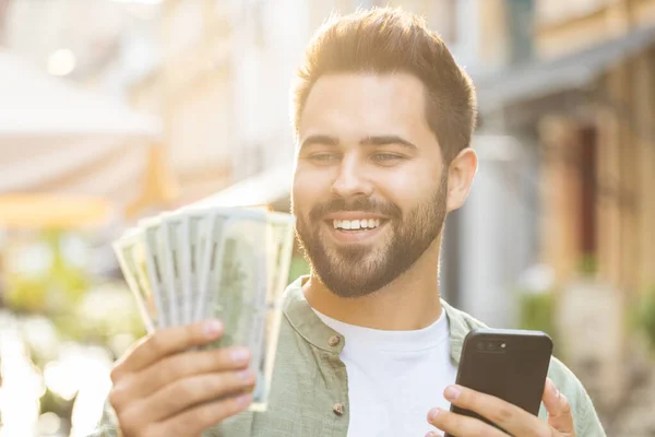 Happy rich young man counting money dollar cash, use smartphone calculator app, plans to order gifts and food delivery online, booking hotel room. Bearded guy tourist in city street. Town outdoors