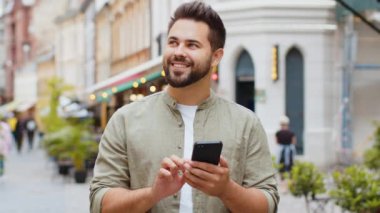Mutlu, gülümseyen sakallı genç adam internet sosyal medya uygulamalarını karıştırarak dışarıda sohbet ederek akıllı telefon yazıları yazıyor. Şehir caddesinde yürüyen bir turist. Kasaba yaşam tarzları