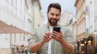 Kafkasyalı genç adam cep telefonuyla kutlama yapıyor. İyi haber kazan, piyango zaferi kazan, açık havada internetten bağış yap. Şehir caddesinde yürüyen mutlu yetişkin erkek turist. Kasaba yaşam tarzları