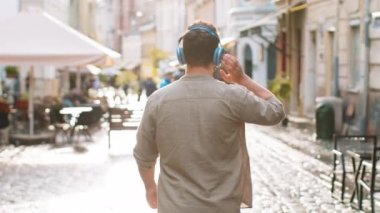 Kablosuz kulaklıklarıyla sokakta yürüyen, enerjik müzik dinleyen, dans eden kameraya dönüp gülümseyen turistin arka görüntüsü. Şehir Günışığı Şehrinde erkek gezginin arka planı