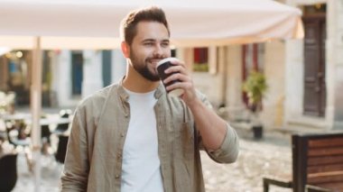 Mutlu sakallı genç adam sabah kahvesinin tadını çıkarıyor sıcak içecek ve dışarıda gülümsüyor. Dinleniyorum, ara veriyorum. Şehirdeki Sunshine Caddesi 'nde yürüyen ve kahve içen bir adam. Dışarıdaki kasaba yaşam tarzları