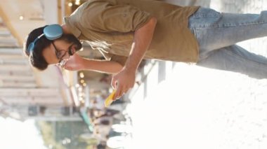 Kablosuz kulaklıklı mutlu, gevşemiş Hintli adam akıllı telefon dansında favori enerjik disko müziğini dinliyor. Turist, Sunshine City caddesinin yanından geçiyor. Dikey
