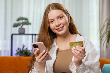 Kredi kartı ve akıllı telefon kullanan genç bir kız para transfer ederken, internetten nakitsiz alışveriş yapar, ev içinde yemek siparişi verir. Mutlu çocuk kanepede oturuyor