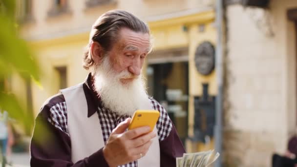 お金のドル現金を持っている幸せな金持ちの老人 スマートフォンの計算機アプリを使用して ギフトを注文する計画 オンライン食品配達 ホテルの部屋を予約します シティストリートの高齢の祖父の観光客 — ストック動画