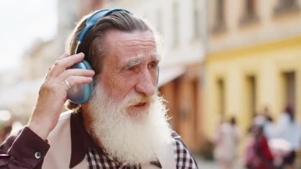 ハッピーリラックスしたワイヤレスヘッドフォンで最高のひげを被った老人を選んで お気に入りのエネルギッシュディスコロックNロールミュージックを屋外で踊ります 都市の通りを歩く高齢のおじいちゃん — ストック動画