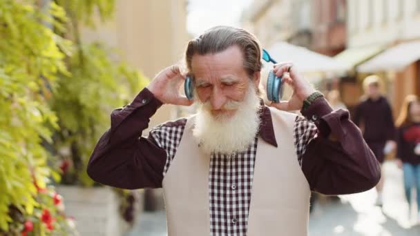 お気に入りのエネルギッシュなディスコロックNロールミュージックを屋外で踊って 選ぶことにハッピーな年老いた男 市街地のカメラを見ているワイヤレスヘッドフォンを離れておじいちゃん — ストック動画