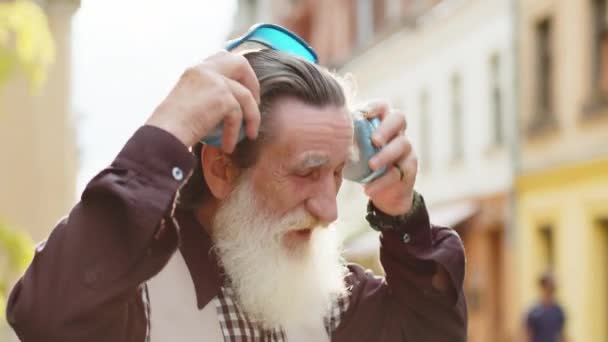 ワイヤレスヘッドフォンを着て 屋外で踊るお気に入りのエネルギッシュディスコロックNロールミュージックを聴くハッピーな老人 都市の通りを歩く成熟した祖父の肖像画 — ストック動画