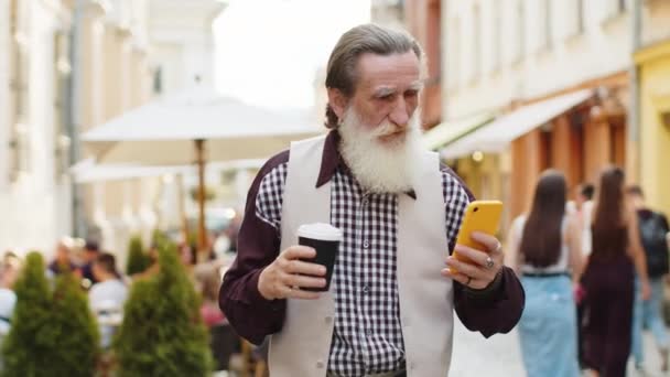 ハッピーシニア老人は インターネットソーシャルメディアWebアプリを閲覧するスマートフォンタイピングテキストメッセージを使用して 朝からコーヒーホットドリンクを楽しんでチャットします ストリートを歩く祖父の観光客 — ストック動画