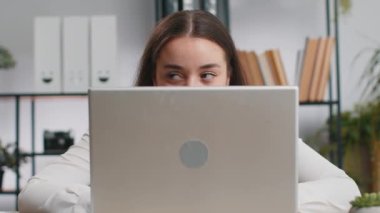 Kendine güvenen iş kadını dizüstü bilgisayarın arkasına saklanıyor, kameraya bakıyor, iş arkadaşlarını gözetliyor, röntgenliyor. Profesyonel serbest çalışan kız kurnaz bakışlarla bilgisayarın arkasından bakıyor.
