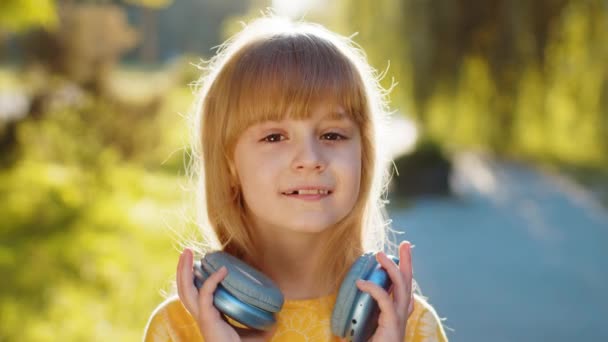 快乐微笑的十几岁以下的孩子们 带着无线耳机选择 在智能手机舞蹈中听着精力充沛的迪斯科音乐 金发女孩看着站在城市日落公园街上的相机 — 图库视频影像