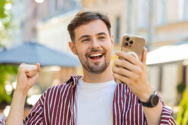 Heyecanlı sakallı adam cep telefonuyla kutlama yapıyor. İyi haberler, başarı, piyango zaferi, açık havada açık havada hediye. Şehir caddesinde yürüyen mutlu beyaz adam. Yaşam Tarzları