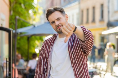 Hoşlanmamak. Üzgün, sakallı ve sakallı bir adam işaret dilini gösteriyor. Hoşnutsuzluğunu, memnuniyetsizliğini, kötü işini, hatasını ifade ediyor. Şehir caddesinde duran hoşnutsuz genç adam.