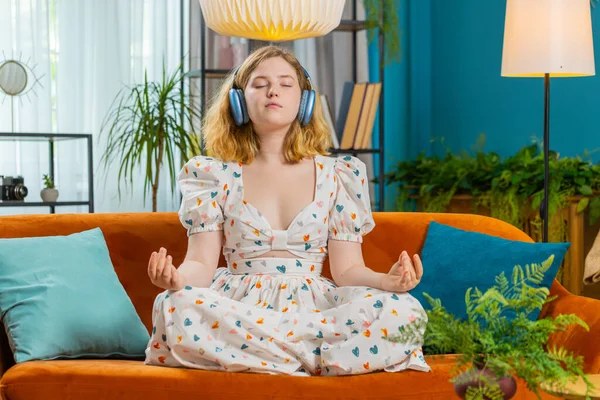 Keep calm down, relax, inner balance. Redhead woman breathes deeply with mudra gesture eyes closed meditating with concentrated thoughts peaceful mind at home apartment. Girl sits on couch. Horizontal