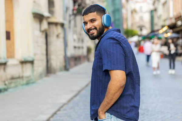 Rear view of indian man tourist in wireless headphones walking through the street outdoors, listening music, dancing, turning to camera and smiling. Back view of guy traveler in urban sunshine city