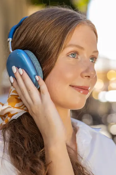 Happy relaxed lovely teenager adult child girl in wireless headphones choosing, listening favorite energetic disco rock n roll music dancing outdoor. Young woman standing in urban city sunny street