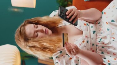 Genç kızıl saçlı kadın kredi kartı ve akıllı telefon kullanarak para transfer ediyor, internetten nakit para almıyor, evde yemek siparişi veriyor. Mutlu kız kanepede oturuyor. Dikey