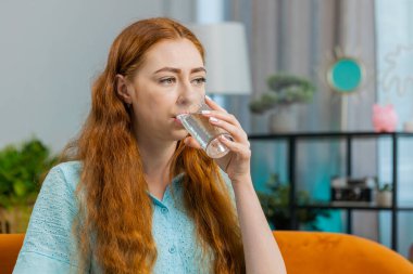 Susamış kızıl saçlı kadın elinde bir bardak su tutarak susuzluğu gidermek için bir yudum su içiriyor. Oturma odasında oturuyor. İyi yaşam alışkanlıkları, sağlıklı zayıflama, kilo verme konsepti olan bir kız.