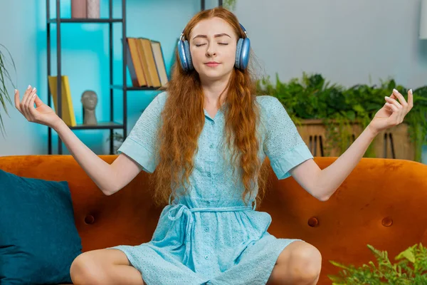Keep calm down, relax, inner balance. Redhead woman breathes deeply with mudra gesture, eyes closed, meditating with concentrated thoughts, peaceful mind at home apartment. Girl sitting on couch