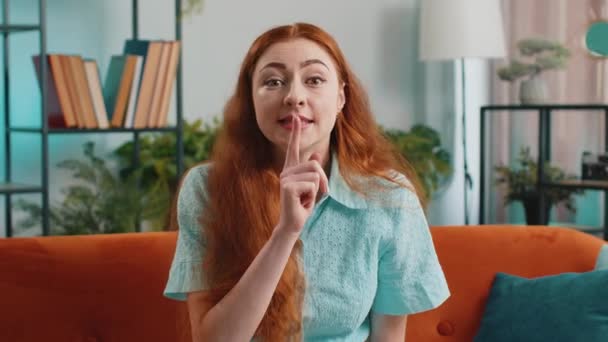 请安静 红头发的年轻女子的肖像把食指压在嘴唇上 使沉默沉默的手势不泄露秘密 现代家庭公寓客厅的女孩坐在沙发上 — 图库视频影像