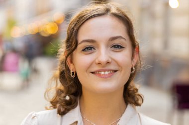Mutlu güzel bir genç kadının portresi. Dostça gülümseyen, kameraya bakan mutlu yüz ifadesi. Hayal kuran, dinlenen, rahatlayan, dışarıda iyi haberler veren. Şehir Güneşi Sokağı arka planında bir kız