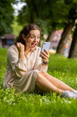 Genç bir kadın cep telefonuyla kutlama yapıyor. İyi haber kazan, piyango kazan, internetten bağış yap. Şehir merkezindeki şehir parkında çimlerde oturan mutlu kız. Yaşam Tarzları