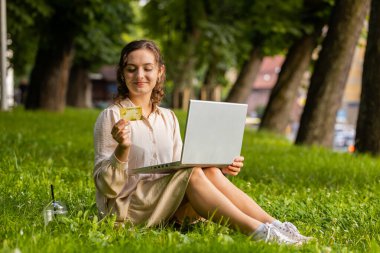 Kredi kartı ve dizüstü bilgisayar kullanan genç bir kadının portresi para transfer ederken, online alışveriş yaparken, yemek siparişi verirken. Şehir parkının arka planında çimlerde oturan kız
