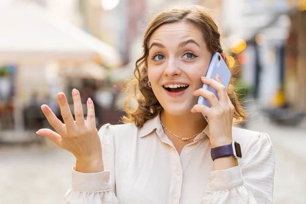 快乐漂亮的女人通过智能手机与人进行远程交流 一个年轻英俊的女孩在电话里说着意想不到的好消息 在城市街道上散步 户外城镇生活方式 — 图库照片