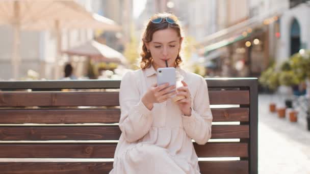 スマートフォンの入力テキスト回答メッセージを使用してかなり若い女性を笑顔モバイル画面のソーシャルメディアアプリを見てオンラインチャット 女の子の観光客は氷と冷たいコーヒーを飲む街の通りのベンチに座っています — ストック動画