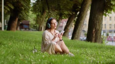 Kablosuz kulaklıklı, neşeli, mutlu genç kadın dışarıda akıllı telefon dansında en sevilen disko rock 'n roll müziğini dinliyor. Şehir parkında çimlerde oturan kız.