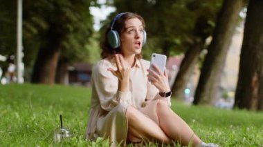 Kablosuz kulaklıklı, neşeli, mutlu genç kadın dışarıda akıllı telefon dansında en sevilen disko rock 'n roll müziğini dinliyor. Şehir parkında çimlerde oturan kız.