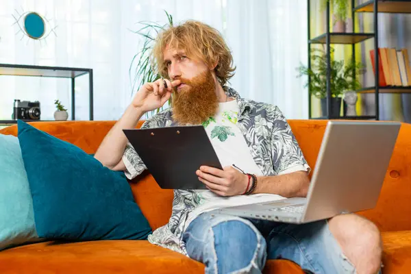 Man freelancer work with documents on home couch, reading information, business papers prepare financial report on laptop. Guy accountant analyzing charts and graph reports documents calculate taxes