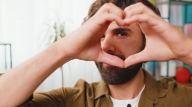 Ben de seni seviyorum. Oturma odasındaki kanepede oturan mutlu Ortadoğulu adam romantik duyguları ifade etmek için kameraya kalp işaretini gösteren bir sembol oluşturuyor. Sadaka. Minnet bağışı.