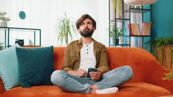 中東の男はソファーに座り 自宅のリビングルームアパートで携帯電話の笑顔を使用しています スマートフォンソーシャルメディアアプリケーションでメッセージコンテンツをオンラインで共有する若者 リラックスした映画を見る — ストック動画