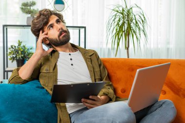 Serbest çalışan bir adam evdeki kanepede dokümanlarla çalışıyor, bilgi okuyor, iş gazeteleri dizüstü bilgisayarda mali rapor hazırlıyor. Haritaları analiz eden muhasebeci ve grafik raporları. Vergileri hesaplayan belgeler.