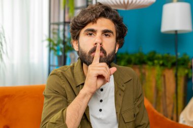 Shh be quiet please. Middle eastern man presses index finger to lips makes silence hush gesture sign do not tells gossip secret. Bearded arabian guy at home apartment living room sitting on couch clipart