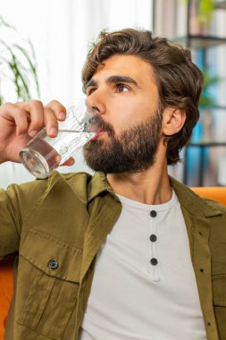 Susamış adamın elinde bir bardak doğal filtrelenmiş su var. Oturma odasında susuz kalmasın diye bir bardak su içiriyor. Ters osmoz filtresinden sonra su. İyi yaşam alışkanlıkları olan, sağlıklı, kilo vermiş bir adam.