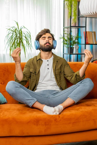 Keep calm down, relax. Middle eastern arabian man listening music breathes deeply, eyes closed meditating with concentrated thoughts, peaceful mind. Bearded guy sits at home in room on couch. Vertical
