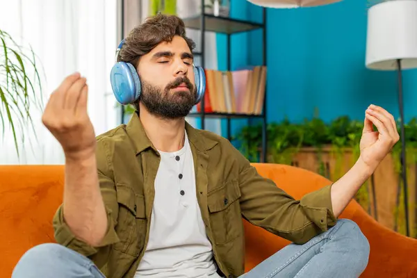 Keep calm down, relax. Middle eastern arabian man listening music breathes deeply, eyes closed meditating with concentrated thoughts, peaceful mind. Young bearded guy sits at home living room on couch