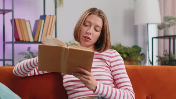 年轻的黑发女人放松有趣的书翻页笑着欣赏文学在舒适的沙发上休息 在家里的客厅里 一个心平气和 开朗的姑娘坐在沙发上 — 图库视频影像