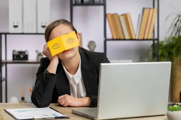 Inefficient tired business woman working sleeping on laptop computer with eyes stickers on face at office workplace desk. Caucasian lazy manager freelancer girl. Business people cheating to sleep