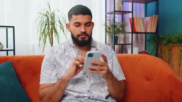 ソファーに座っているインディアン男性は 家庭のリビングルームアパートで携帯電話の笑顔を使用しています スマートフォンソーシャルメディアアプリケーションでメッセージのコンテンツをオンラインで共有する若いアラビアの男 リラックスした映画を見る — ストック動画