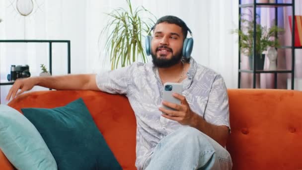 在家里沙发上跳舞的印度男人在无线耳机里开心地放松着 在智能手机娱乐节目中选择了听着最喜欢的精力充沛的迪斯科摇滚音乐 人们周末活动 — 图库视频影像