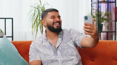 Mutlu Hintli blog yazarı akıllı telefondan selfie çekiyor, abonelerle online video görüşmesi yapıyor, sosyal medya için hikayeler kaydediyor. Modern ev dairesindeki adam kanepede oturuyor.