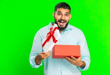 Mutlu Hintli adam kırmızı kurdeleli doğum günü hediyesi kutusunu açıyor. Tatil sürprizi konsepti. Gülümseyen Arap sakallı adam yeşil renkli arka planda izole edilmiş harika bir kutlama hediyesi aldı.