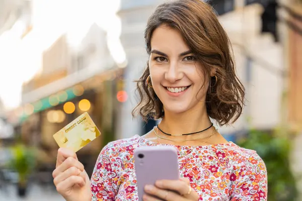 Young woman using credit bank card smartphone while transferring money, purchases online shopping, order food delivery, booking hotel room. Smiling girl standing in urban city sunny street outdoors